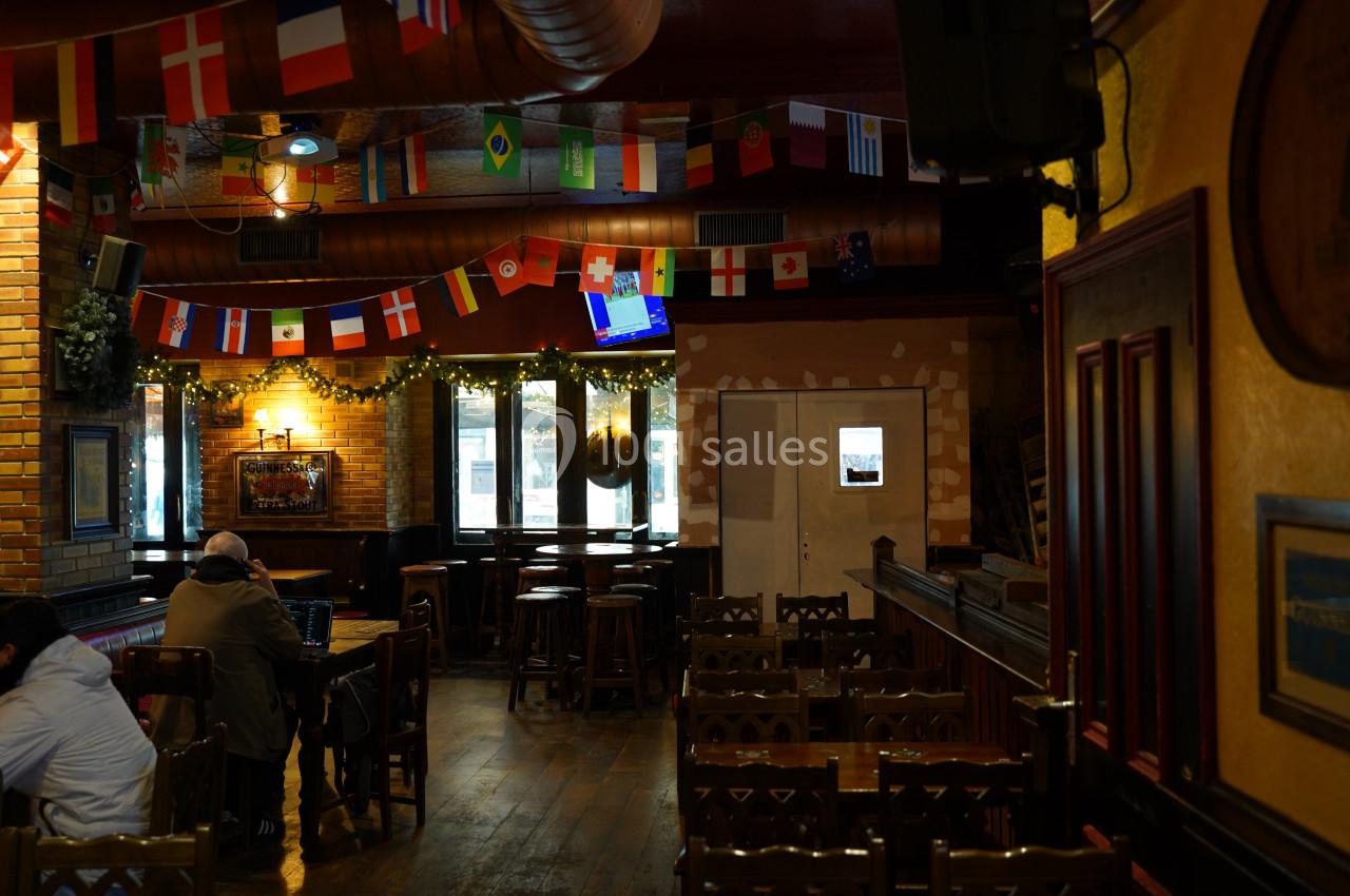Salle de bar chaleureuse avec des drapeaux suspendus, des tables en bois et un écran diffusant une émission.