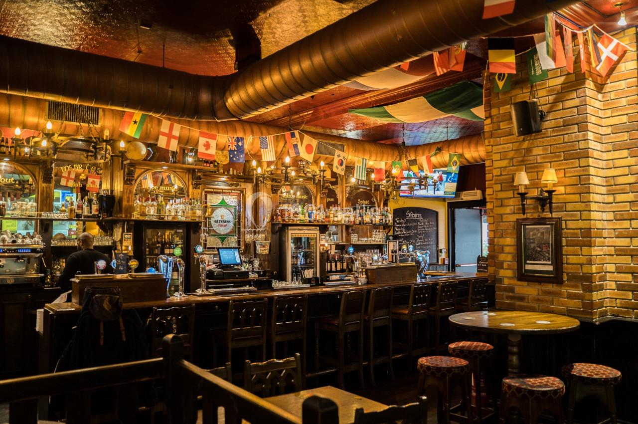 Intérieur d'un pub chaleureux avec un bar en bois, des drapeaux suspendus et des tables entourées de chaises.