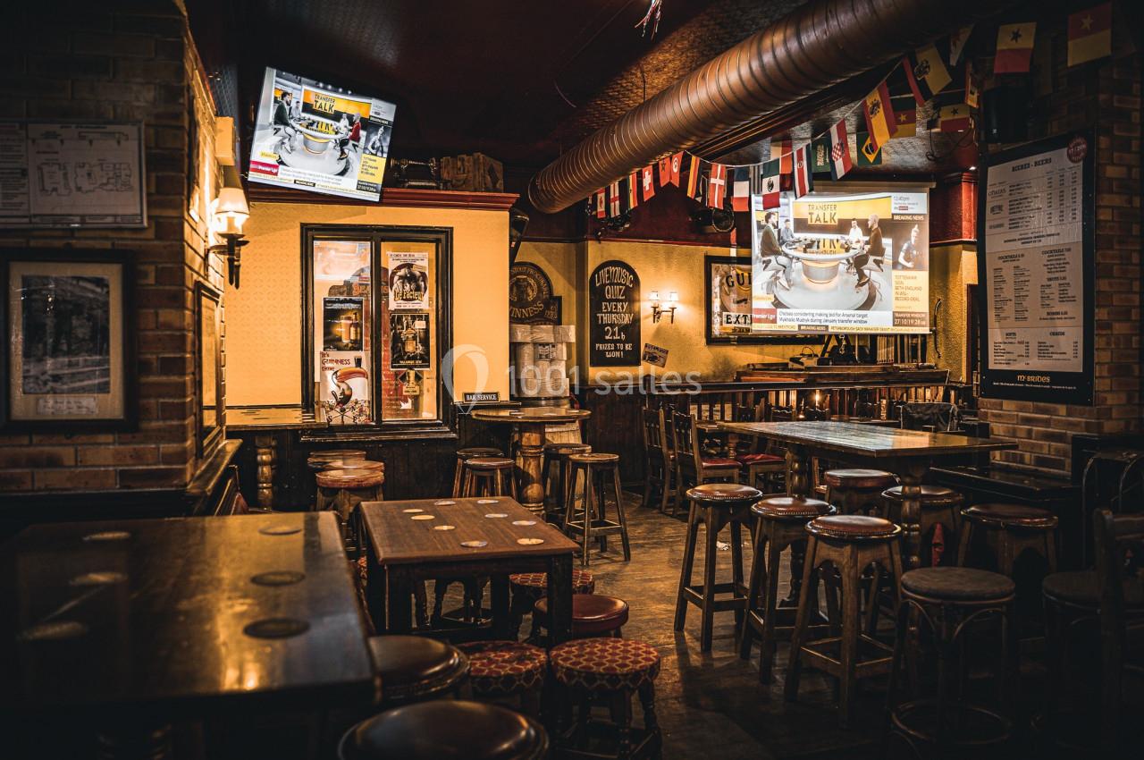 Intérieur d'un bar chaleureux avec tables en bois, tabourets hauts, écrans allumés et décorations de drapeaux.