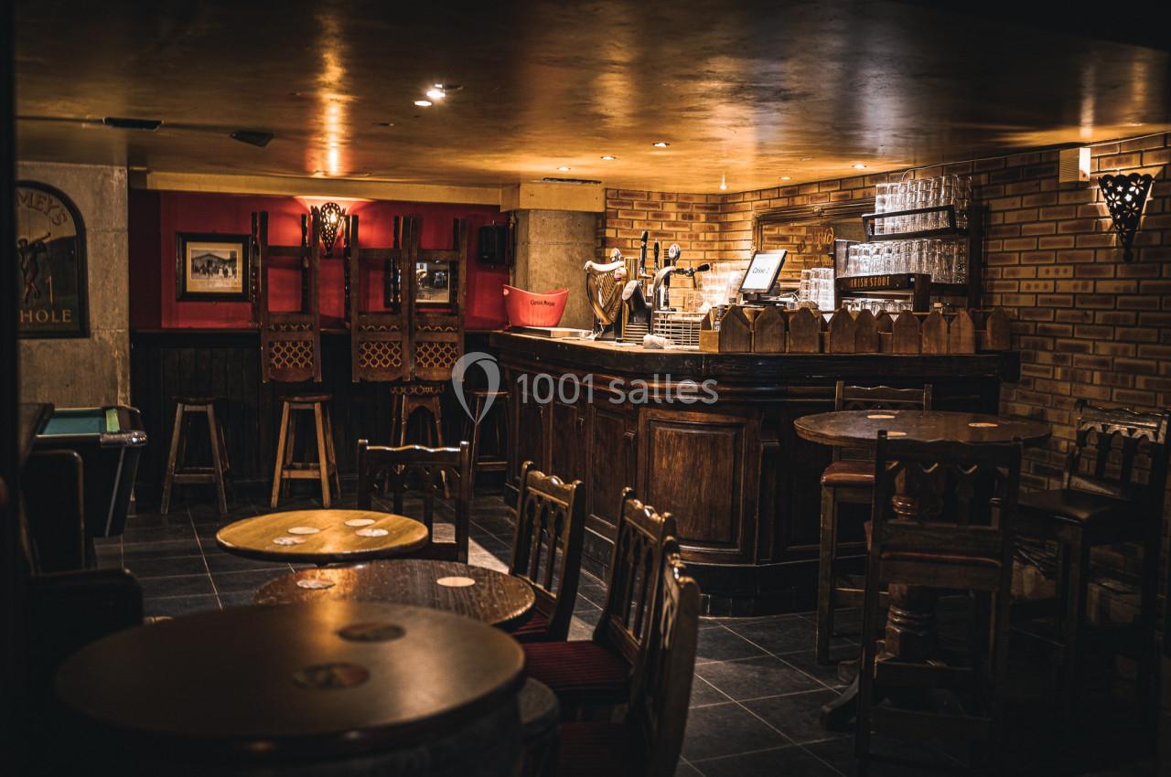 Intérieur d'un bar chaleureux avec comptoir en bois, tabourets hauts, tables rondes et éclairage tamisé.