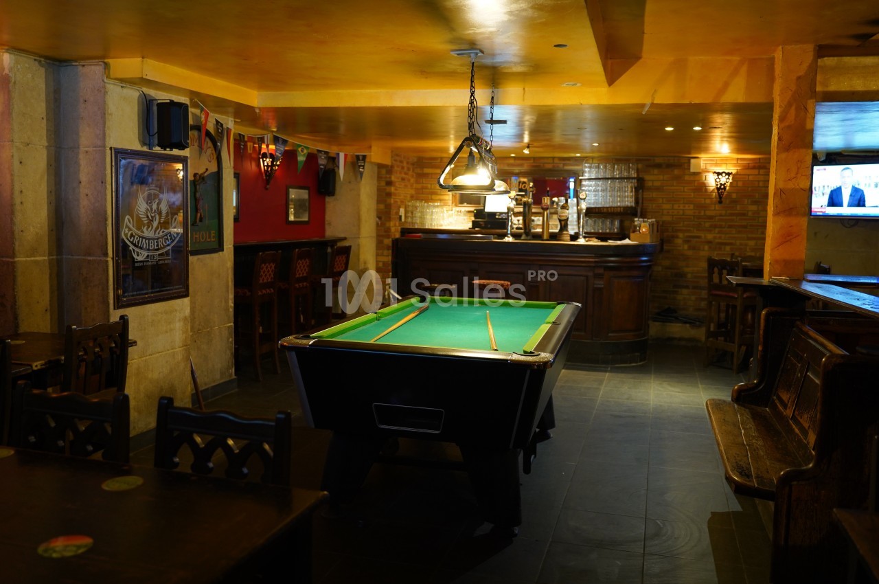 Salle de bar avec une table de billard au centre, un comptoir en bois au fond et des chaises disposées autour.