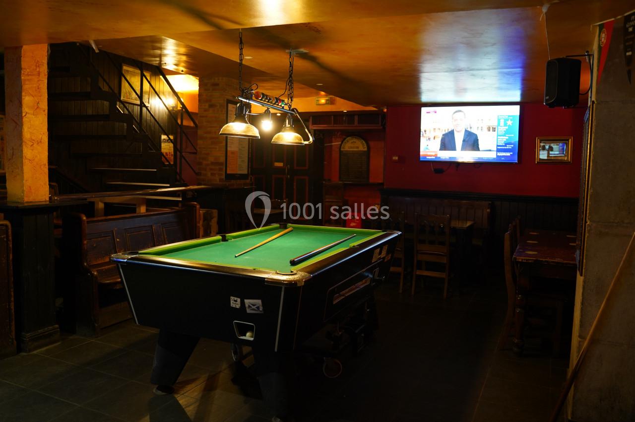 Table de billard éclairée dans un bar à l'ambiance tamisée, avec un écran de télévision allumé en arrière-plan.