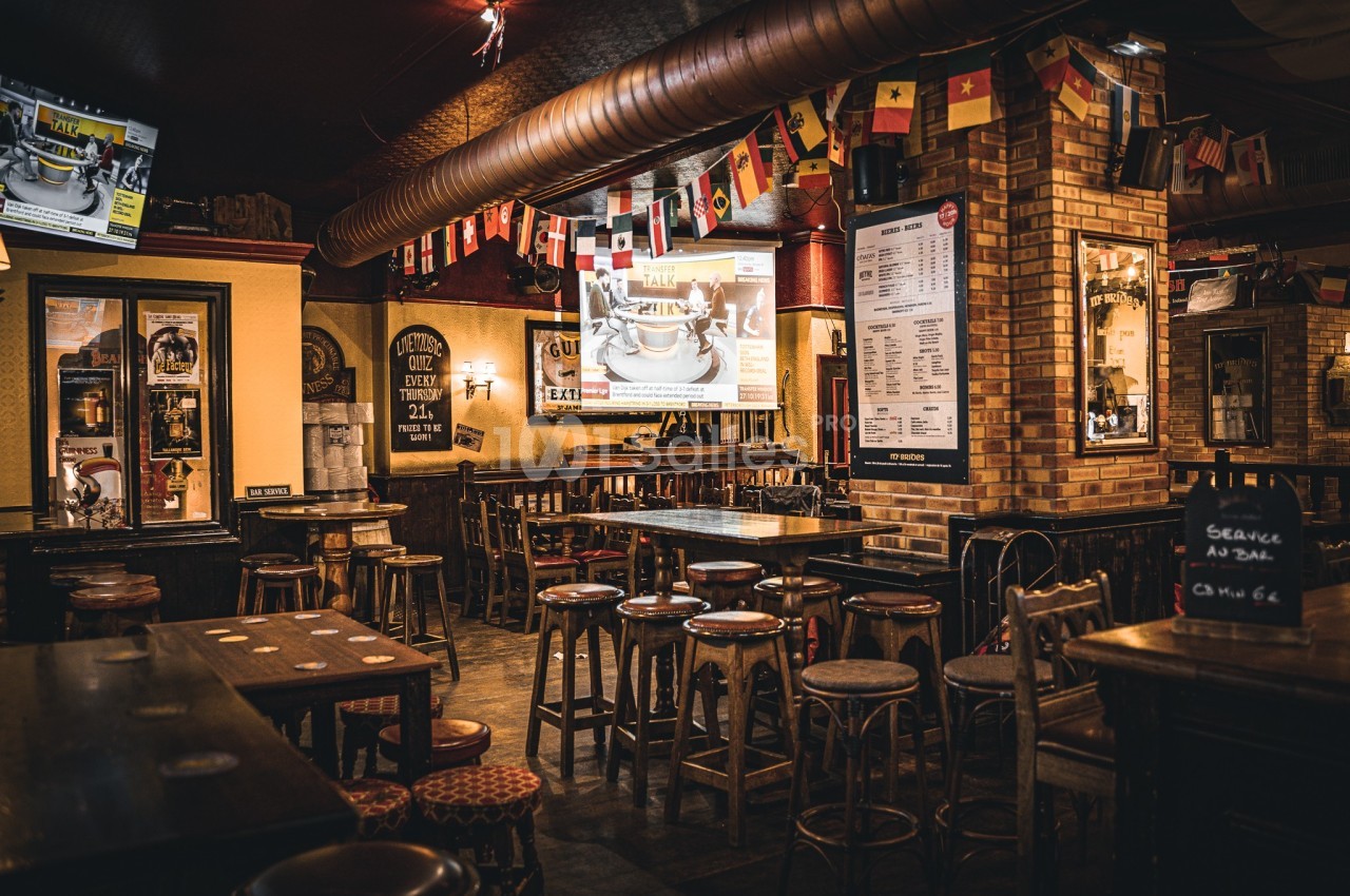 Intérieur d'un bar avec des tables en bois, des tabourets, des drapeaux suspendus et un écran affichant une image.