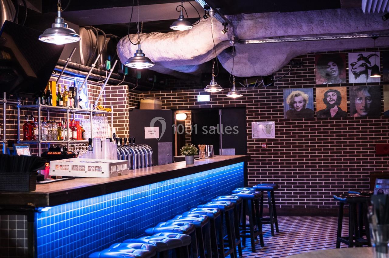 Bar intérieur avec comptoir éclairé en bleu, tabourets alignés, murs en briques et décorations murales.