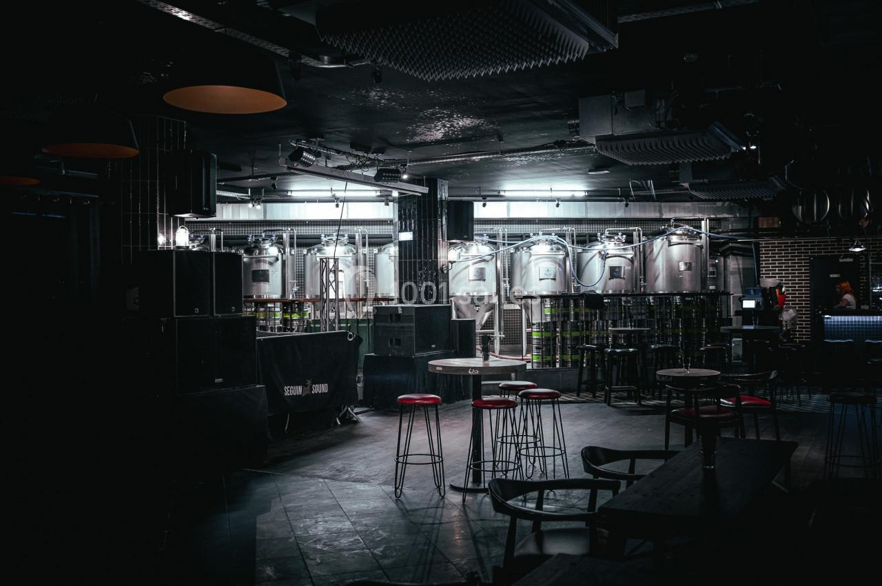 Salle sombre avec des cuves en acier inoxydable, des tabourets rouges et des tables hautes dans un décor industriel.