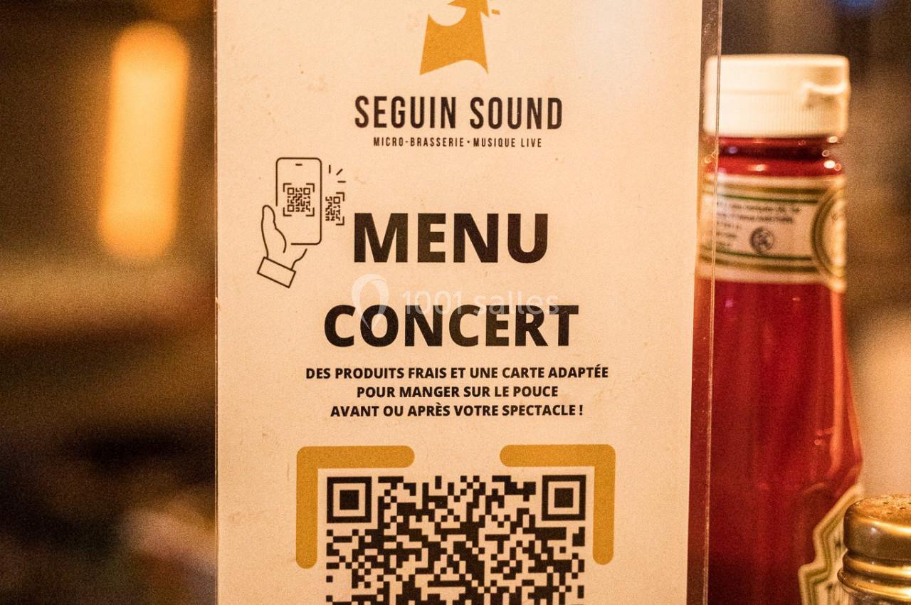 Affiche sur un comptoir présentant un menu concert avec QR code, ketchup et décor flou en arrière-plan.