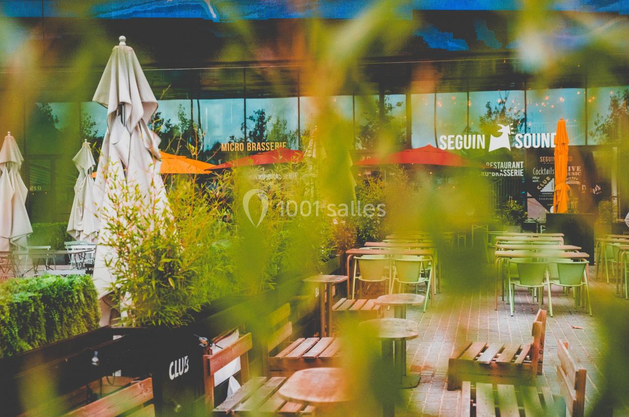 Terrasse extérieure d'un restaurant avec tables, chaises, parasols et végétation, vue à travers des feuillages.