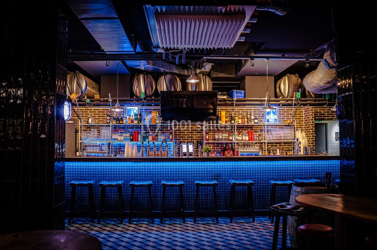 Bar avec comptoir éclairé en bleu, tabourets alignés, étagères remplies de bouteilles et décor industriel en arrière-plan.