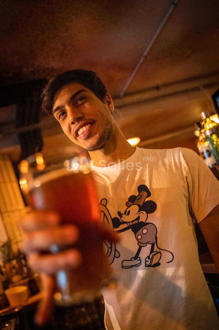 Un homme souriant tend un verre de bière dans un bar à l'éclairage chaleureux.