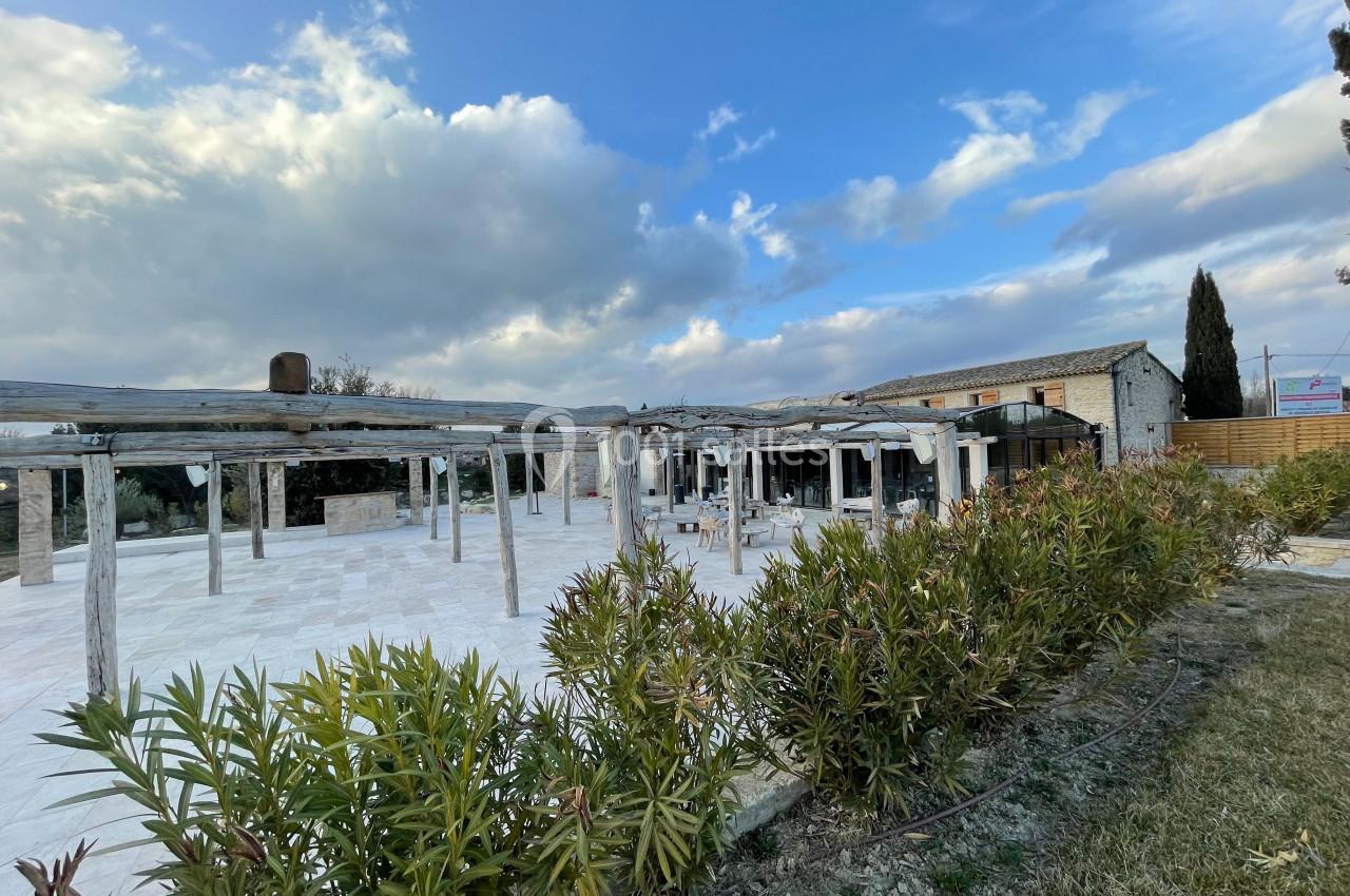 Location salle Aix-en-Provence (Bouches-du-Rhône) - Maneo #12 Cour extérieure avec pergolas en bois, entourée de végétation, devant un bâtiment en pierre sous un ciel partiellement…