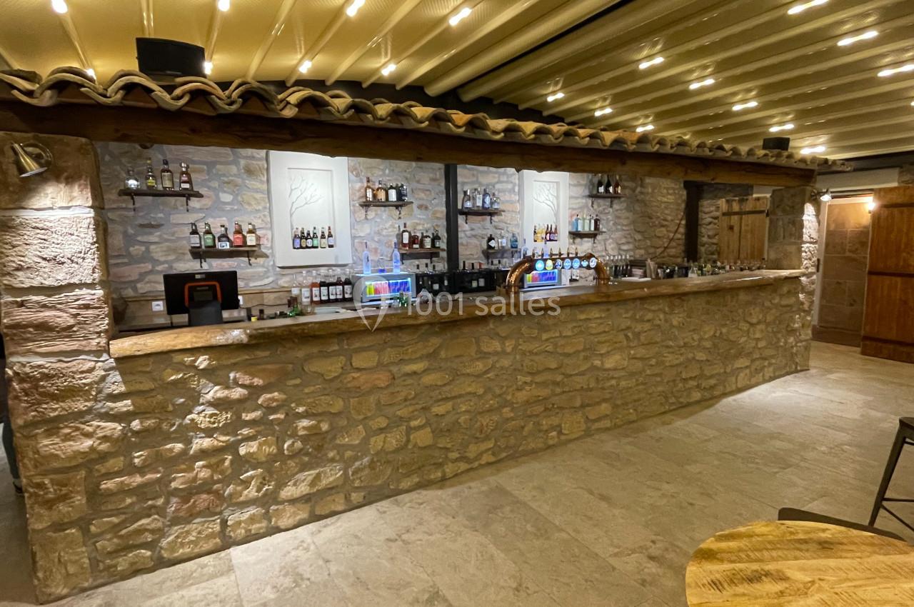 Location salle Aix-en-Provence (Bouches-du-Rhône) - Maneo #13 Comptoir de bar en pierre avec étagères garnies de bouteilles, éclairage chaleureux et plafond à poutres apparentes.
