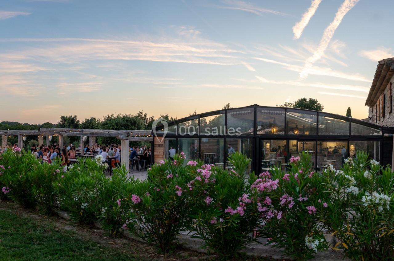 Location salle Aix-en-Provence (Bouches-du-Rhône) - Maneo #6 Bâtisse en pierre avec verrière, terrasse animée et jardin fleuri, sous un ciel au coucher du soleil.