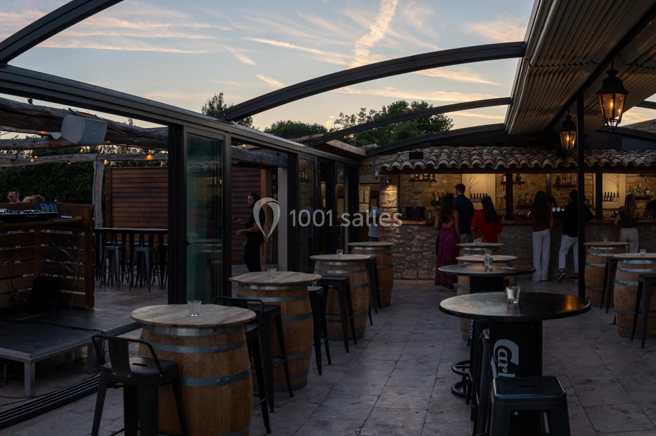 Location salle Aix-en-Provence (Bouches-du-Rhône) - Maneo #7 Terrasse extérieure avec tables en tonneaux, chaises hautes et toit partiellement ouvert, éclairée par une lumière douce.