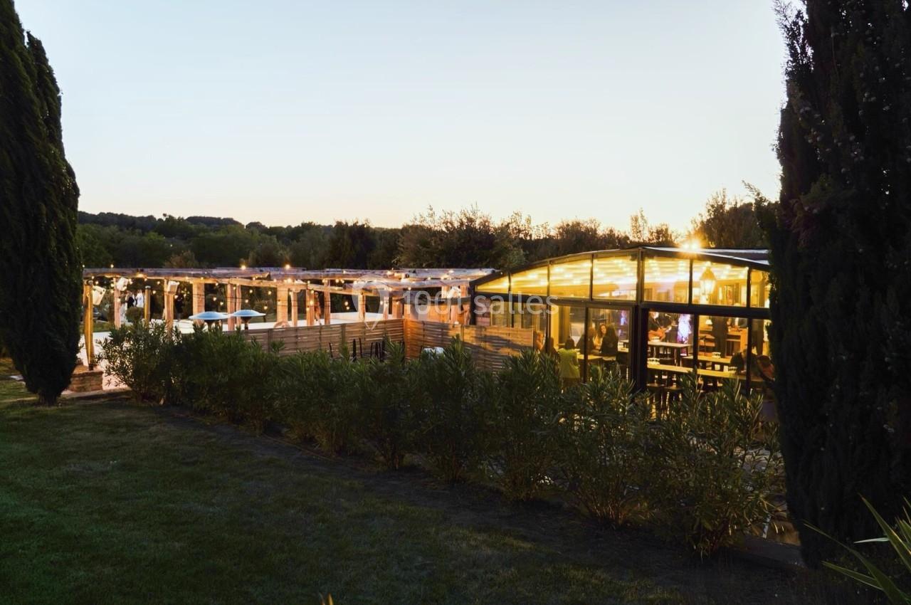 Location salle Aix-en-Provence (Bouches-du-Rhône) - Maneo #17 Vue d'un restaurant éclairé en soirée, entouré de végétation et d'arbres, avec une terrasse illuminée.