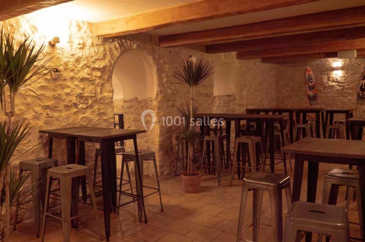 Location salle Aix-en-Provence (Bouches-du-Rhône) - Maneo #18 Salle avec murs en pierre, tables hautes en bois, tabourets métalliques et plantes décoratives sous un éclairage chaleureux.