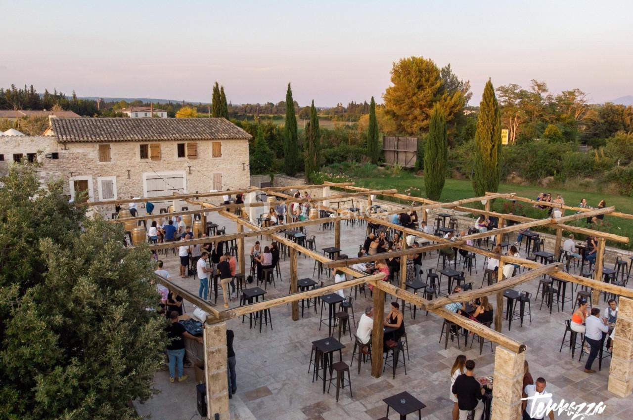 Vue aérienne d'une terrasse en pierre avec des tables hautes, des invités debout et un bâtiment en arrière-plan.