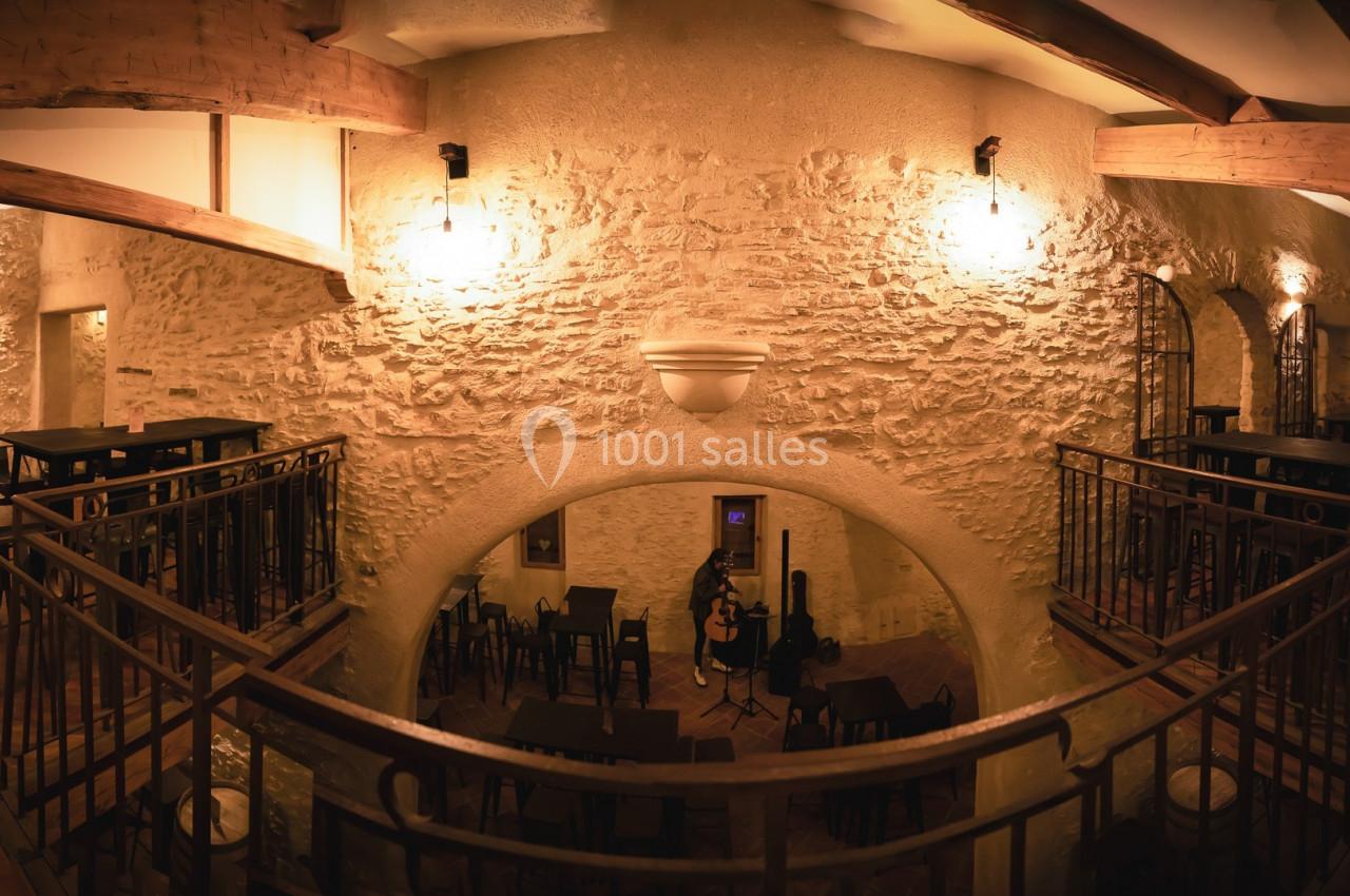 Salle intérieure en pierre avec mezzanine, éclairage chaleureux, tables hautes et chaises disposées autour.