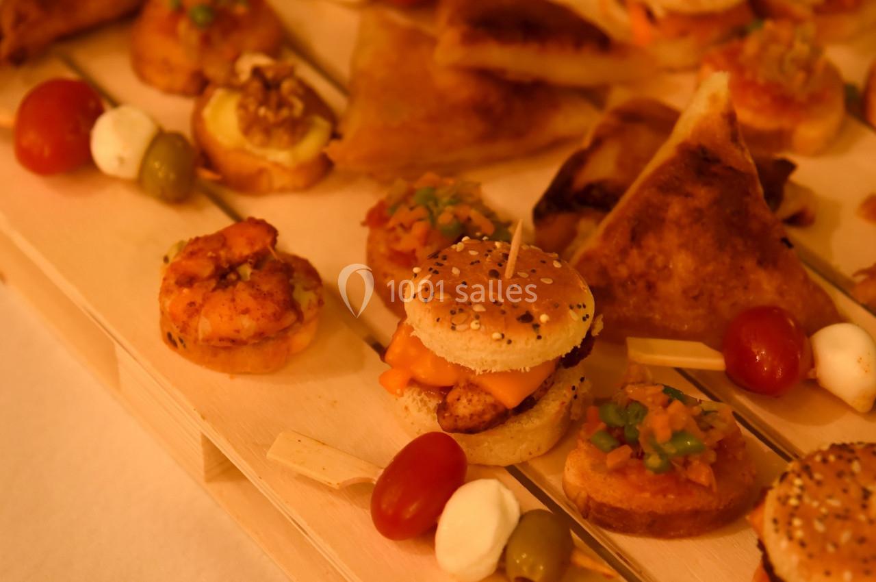 Plateau en bois garni de bouchées apéritives variées, incluant mini-burgers, samoussas, olives et tomates cerises.