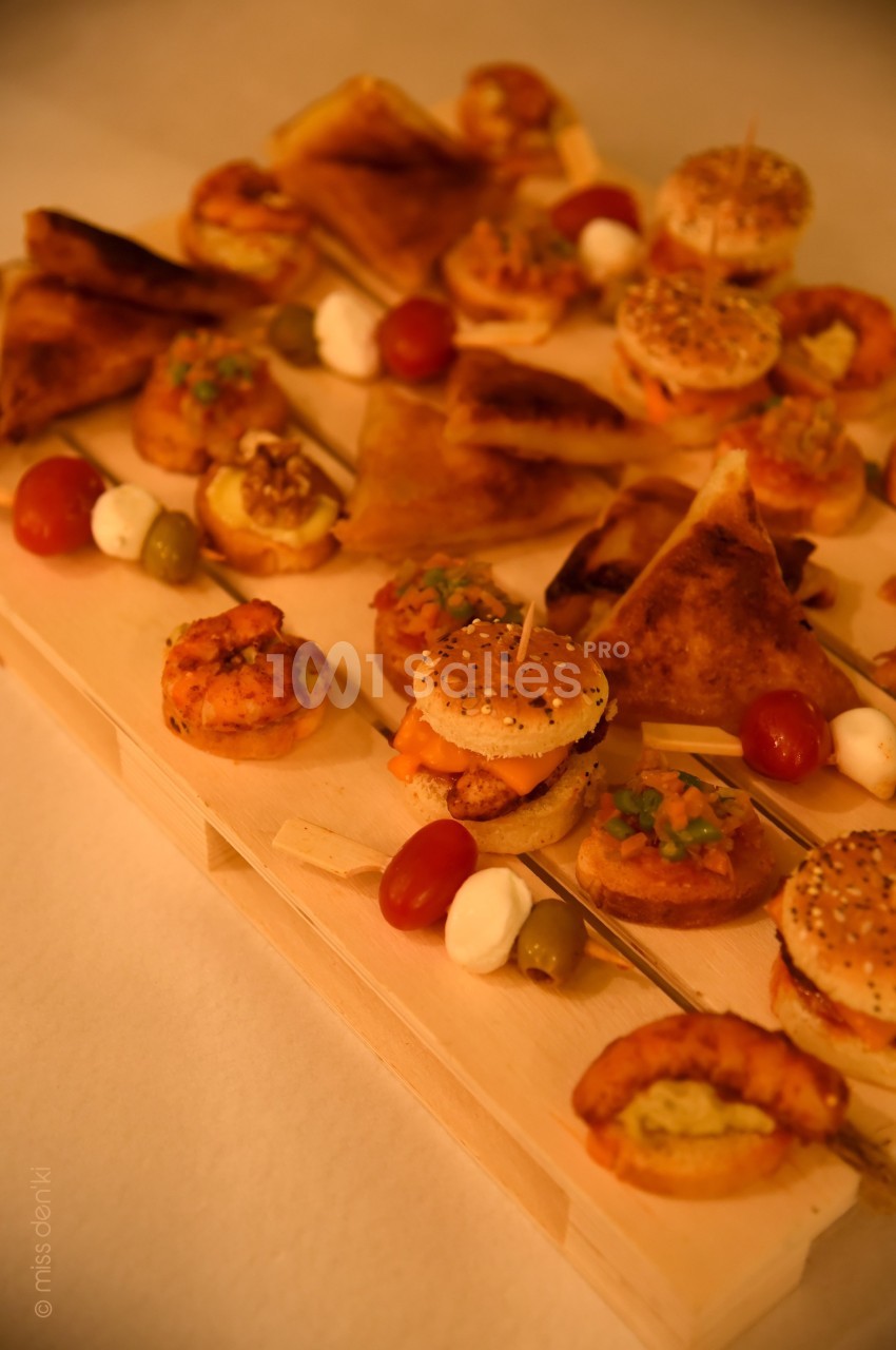 Assortiment de bouchées apéritives variées disposées sur un plateau en bois, incluant mini-burgers, feuilletés et brochettes.