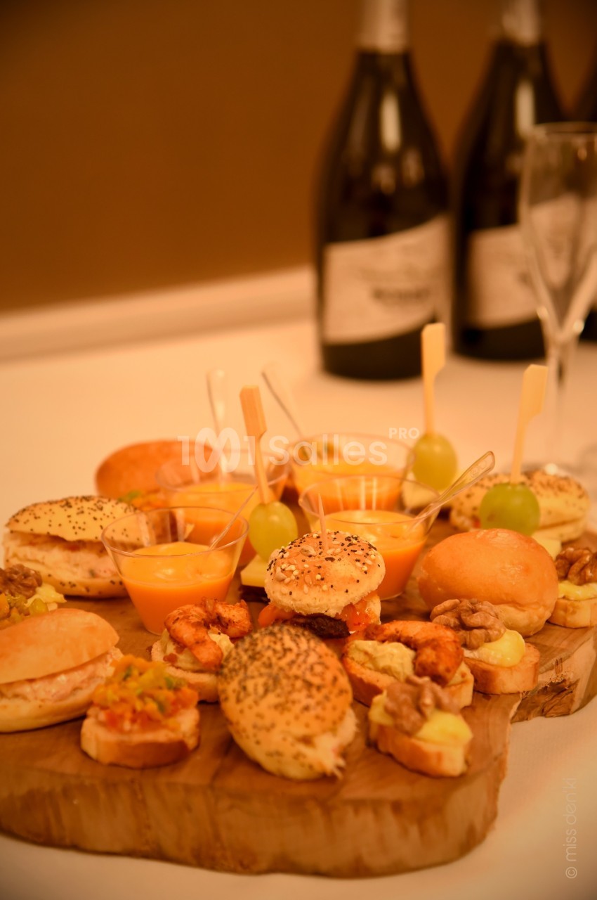 Plateau en bois avec amuse-bouches variés, mini-sandwichs, verrines et brochettes, deux bouteilles en arrière-plan.