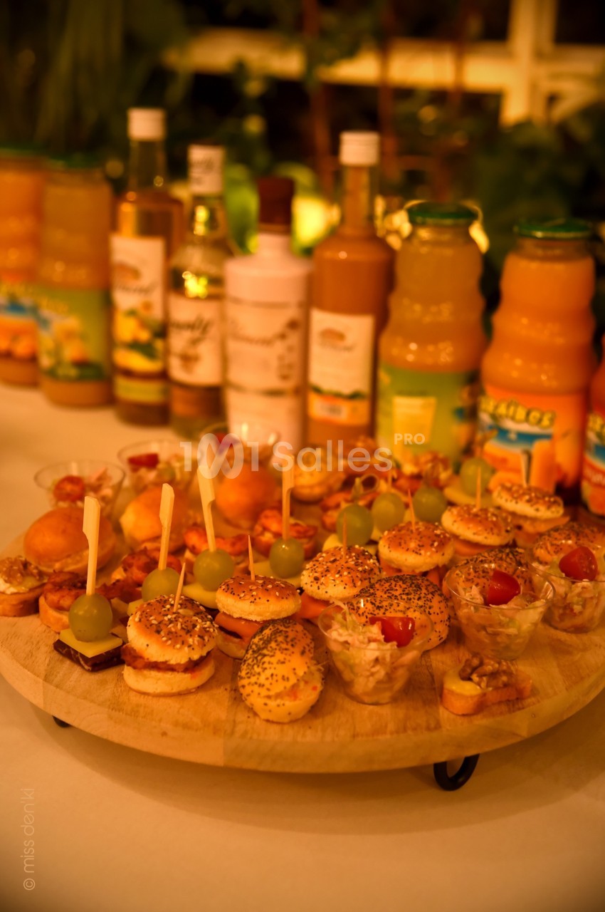 Plateau de mini-burgers, verrines et amuse-bouches disposés devant des bouteilles de jus de fruits variés.