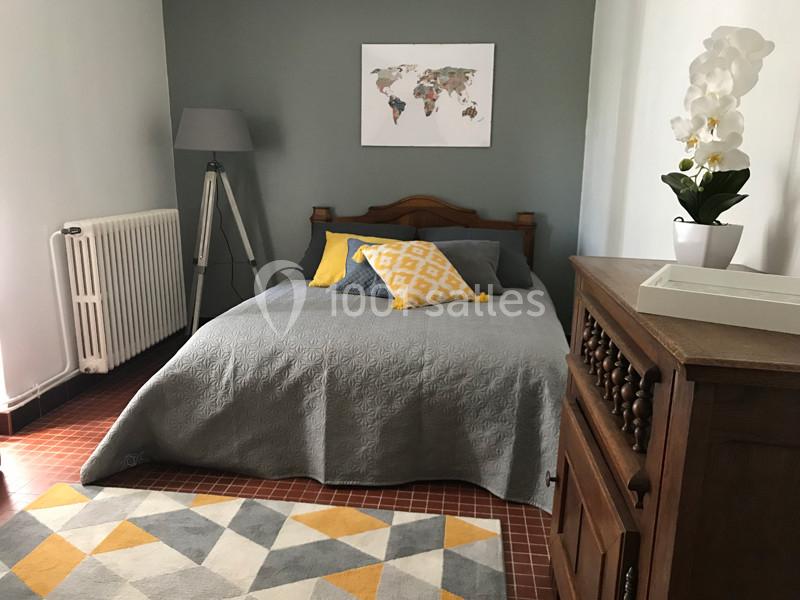 Chambre avec lit double, couvre-lit gris, coussins jaunes, lampe sur pied, carte du monde au mur et orchidée blanche.