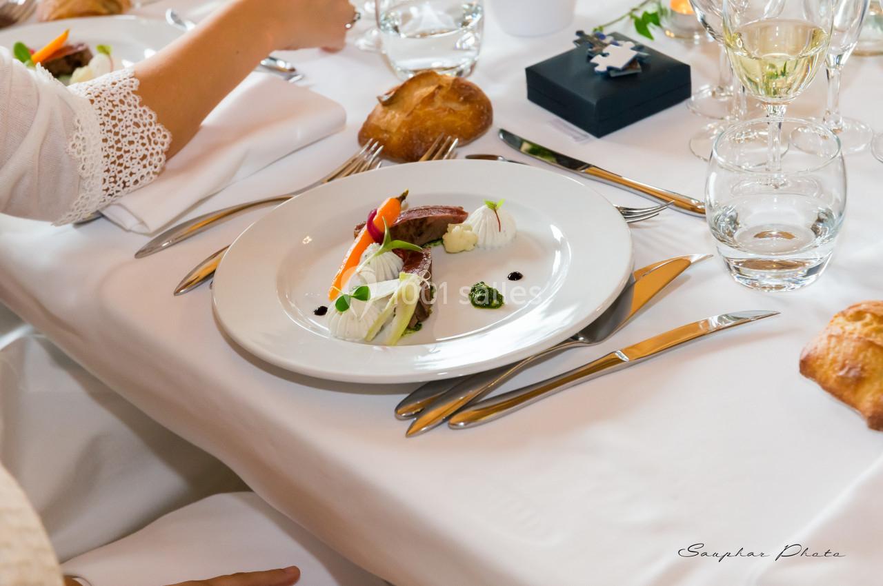 Assiette élégamment dressée avec légumes, viande et sauce, sur une table blanche avec couverts et verres.