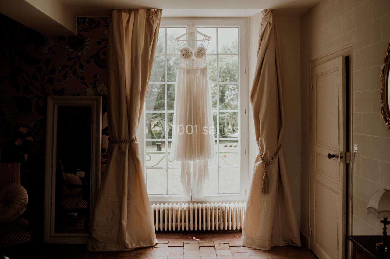 Robe de mariée suspendue devant une grande fenêtre, encadrée par des rideaux beiges dans une pièce lumineuse.