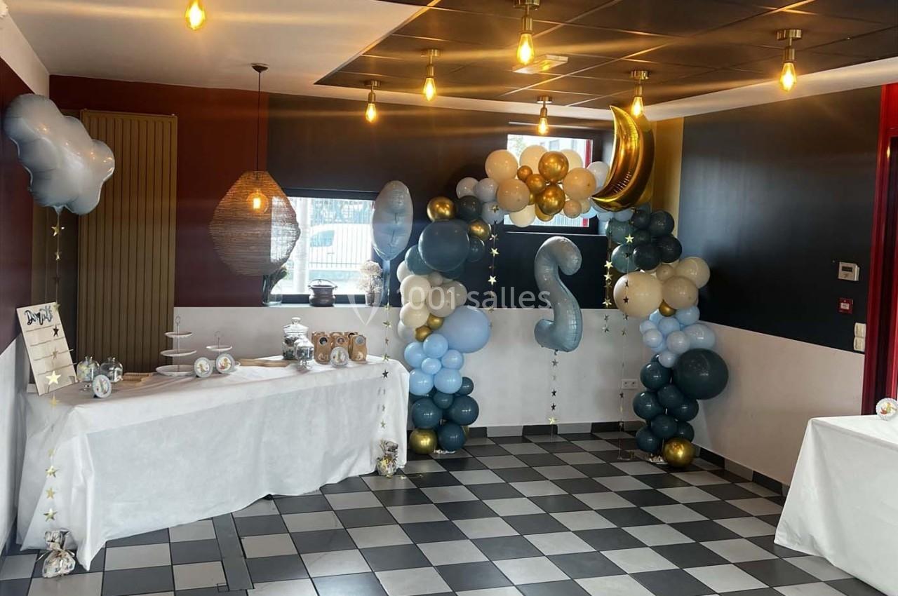 Décoration de salle avec ballons bleus, blancs et dorés formant une arche, tables dressées et éclairage suspendu.