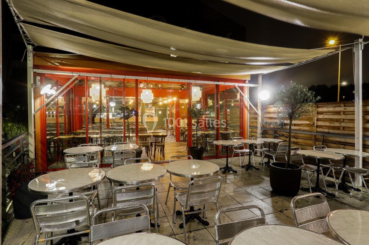 Terrasse extérieure d'un restaurant avec tables et chaises en métal, éclairée par des lampes sous des toiles tendues.