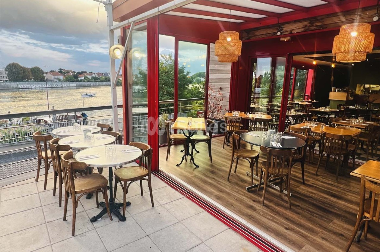 Terrasse et salle intérieure d'un restaurant avec vue sur une rivière, mobilier en bois et ambiance chaleureuse.