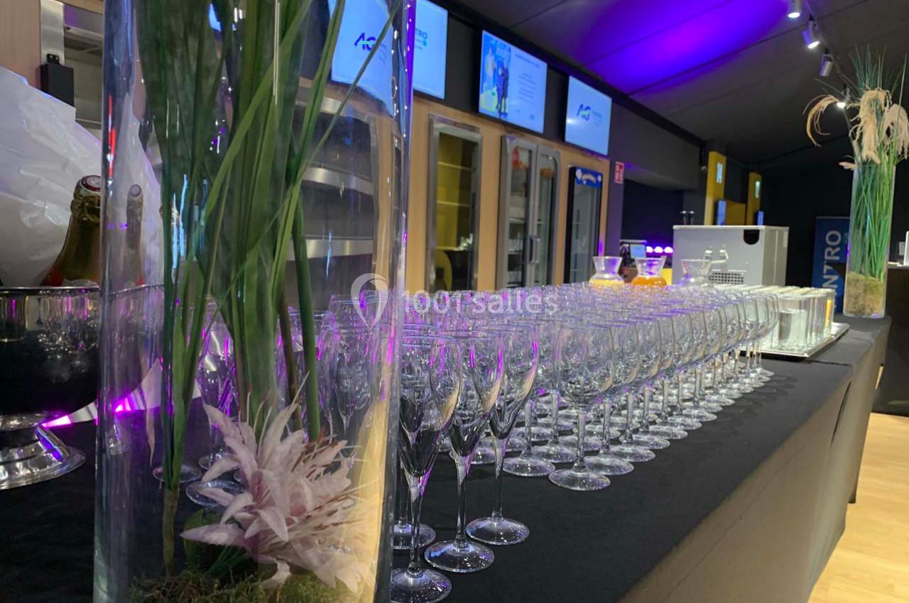 Rangée de verres à vin alignés sur une table noire, avec des vases décoratifs et un éclairage violet en arrière-plan.