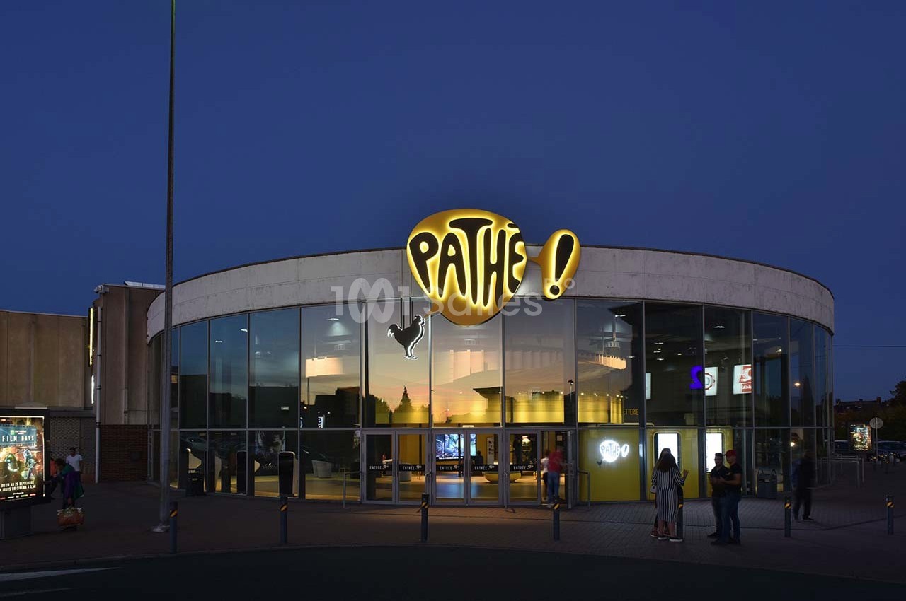 Façade d'un cinéma Pathé éclairée en soirée, avec des passants devant l'entrée vitrée.