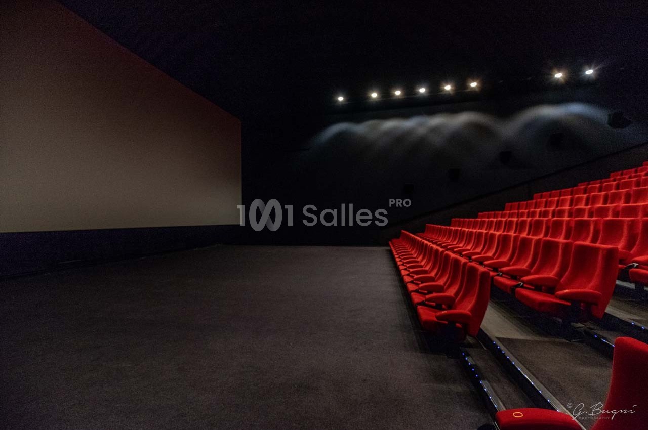 Salle de cinéma vide avec rangées de sièges rouges et un grand écran éteint dans une ambiance tamisée.