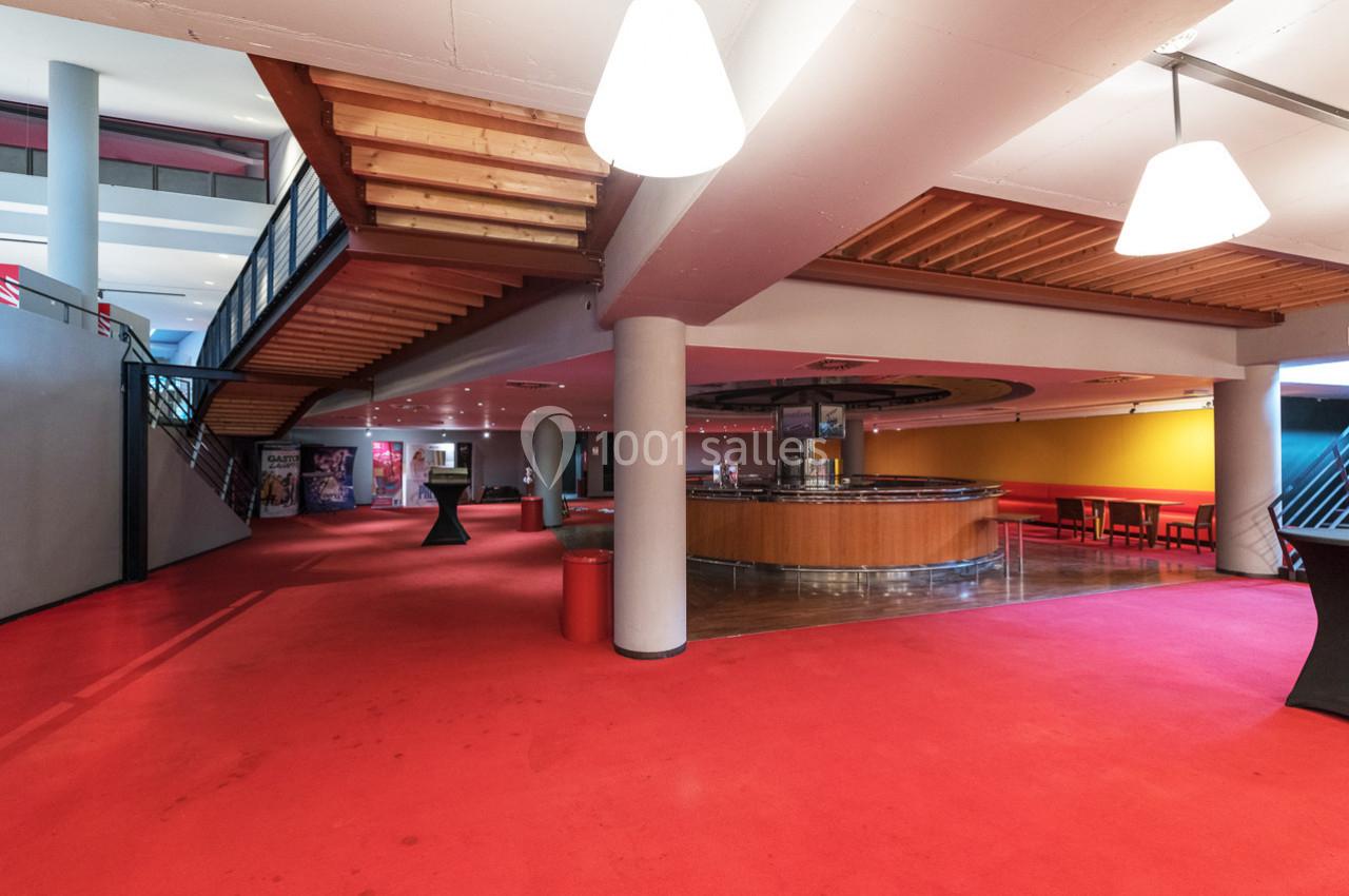 Espace intérieur avec moquette rouge, escalier en bois, comptoir circulaire et tables dans un coin lumineux.