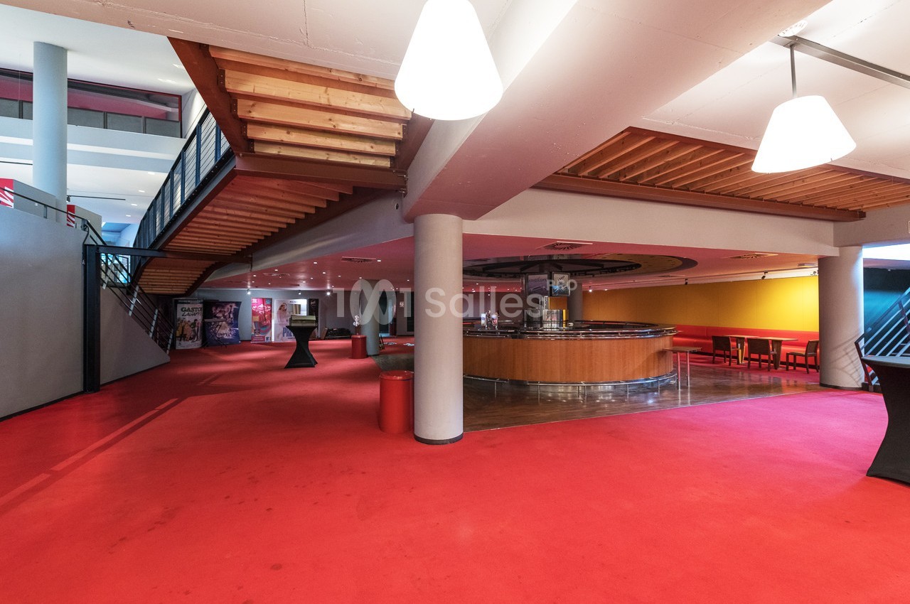 Espace intérieur avec moquette rouge, escalier en bois, comptoir circulaire et tables dans un coin lumineux.