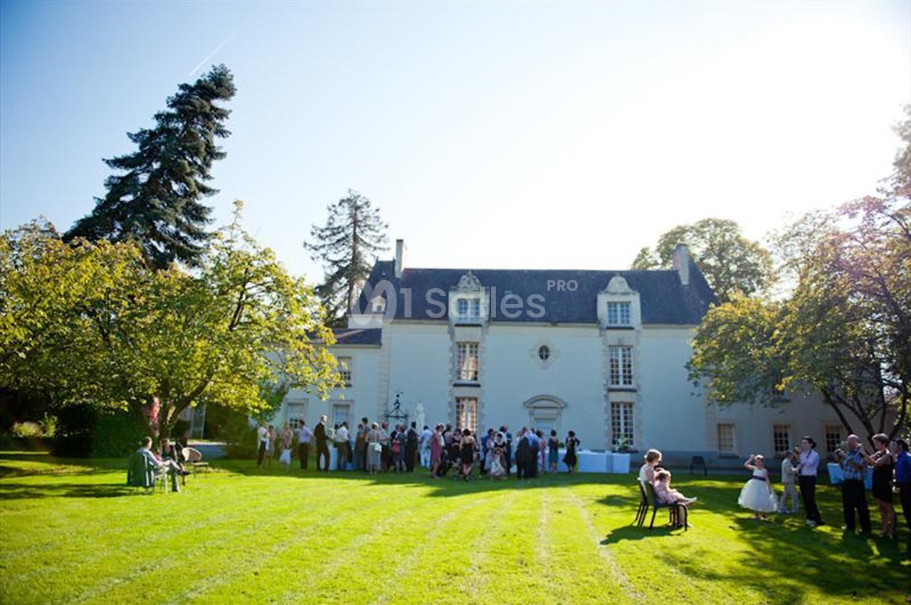 Groupe de personnes rassemblées devant un manoir blanc entouré d'un grand jardin verdoyant par une journée ensoleillée.