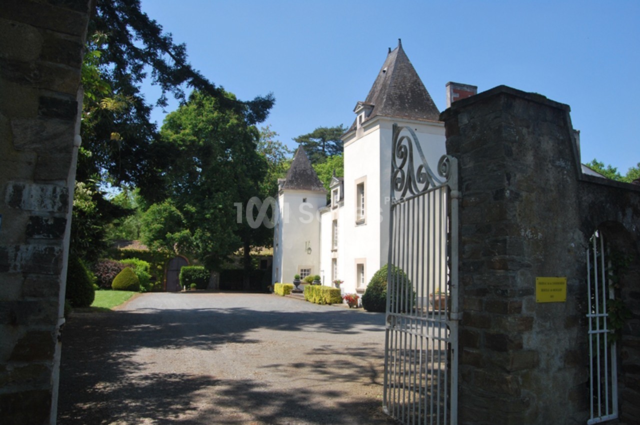 Entrée d'une propriété avec portail ouvert, allée gravillonnée, bâtiment blanc à tourelles et jardin arboré.