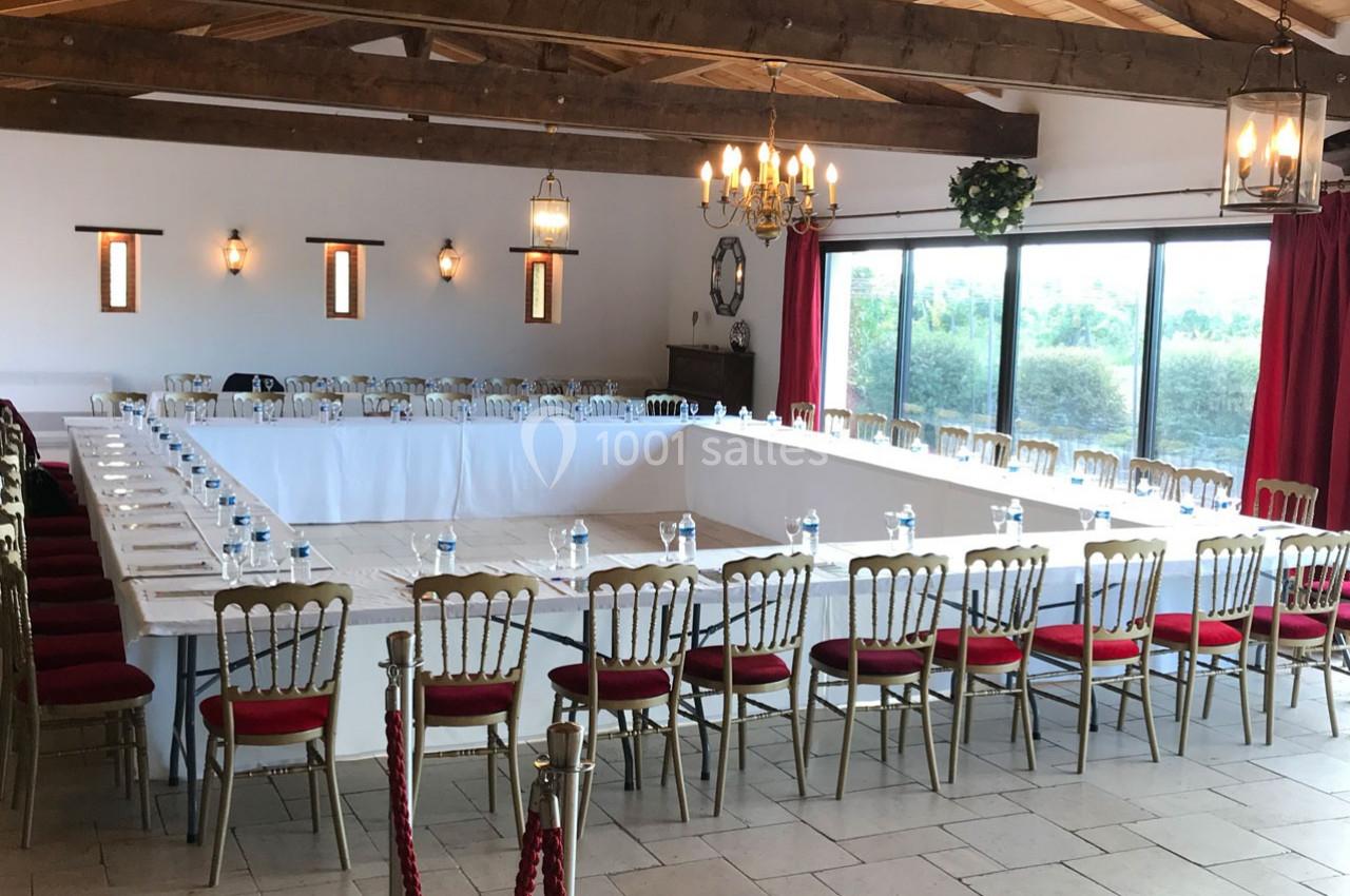 Salle de réunion lumineuse avec tables disposées en U, chaises rouges, grandes fenêtres et poutres apparentes.