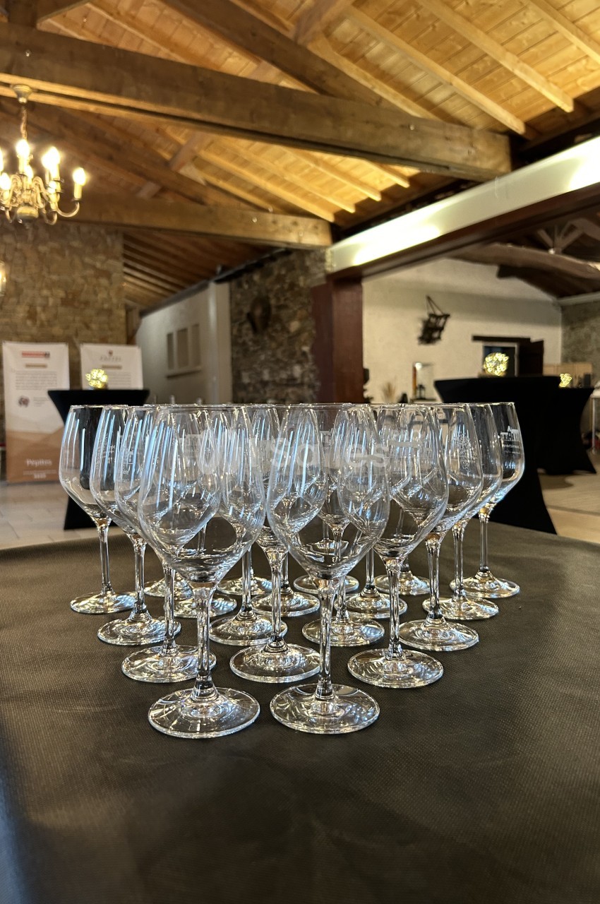 Rangée de verres à vin vides alignés sur une table dans une salle avec poutres apparentes et murs en pierre.