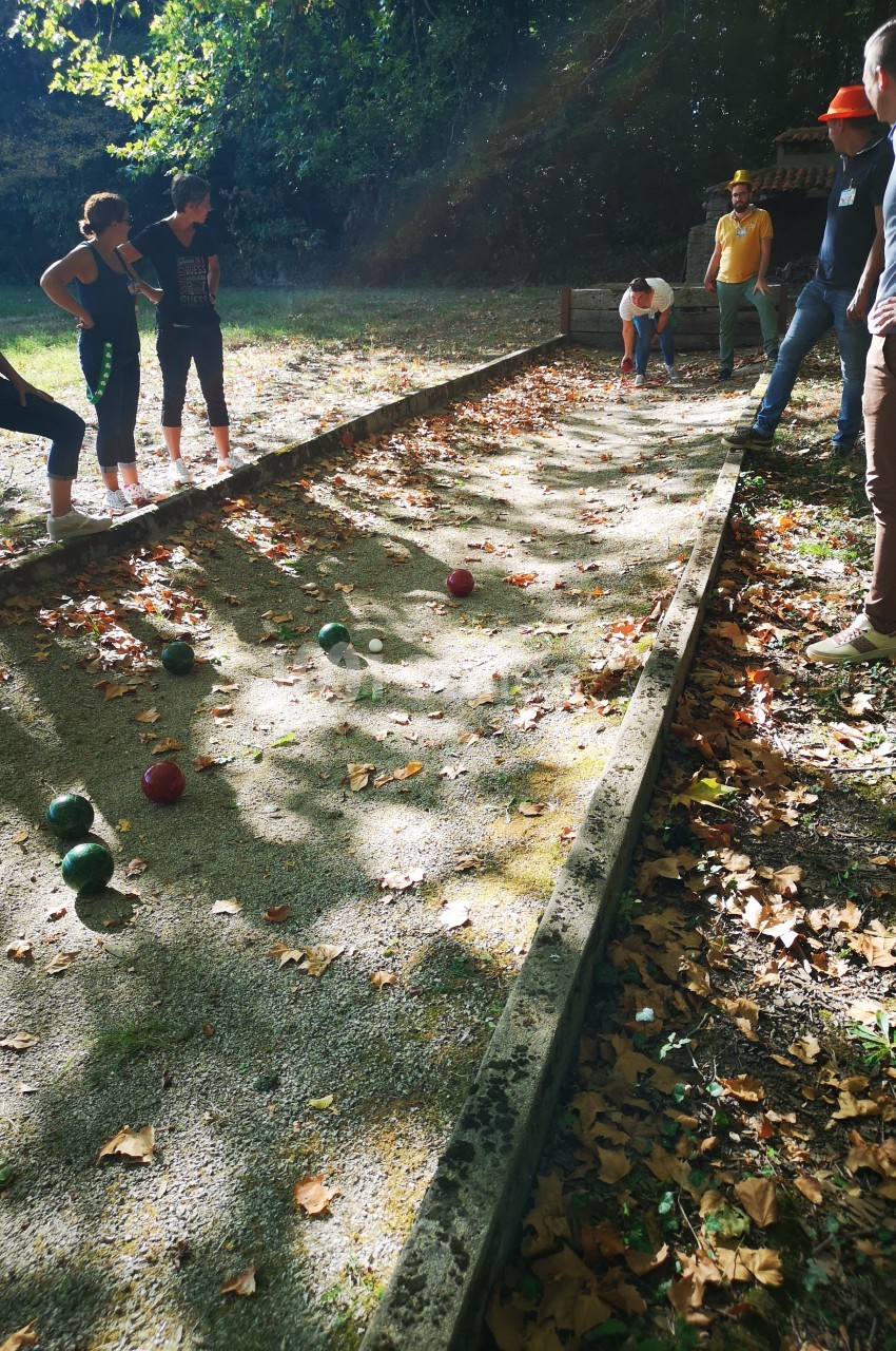 Des personnes jouent à la pétanque sur un terrain ombragé entouré de feuilles mortes.