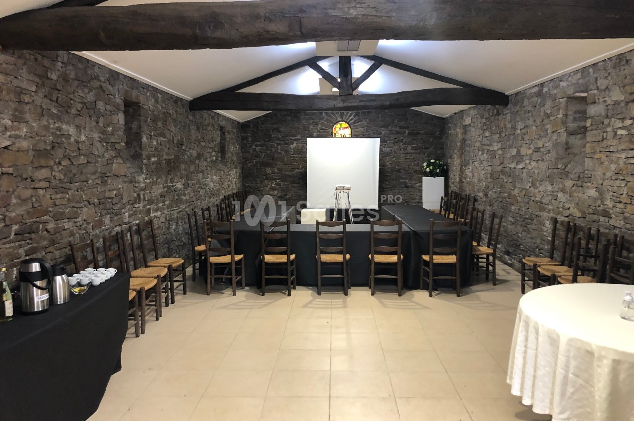 Salle en pierre avec poutres apparentes, tables disposées en U, chaises en bois, écran de projection et table de service.