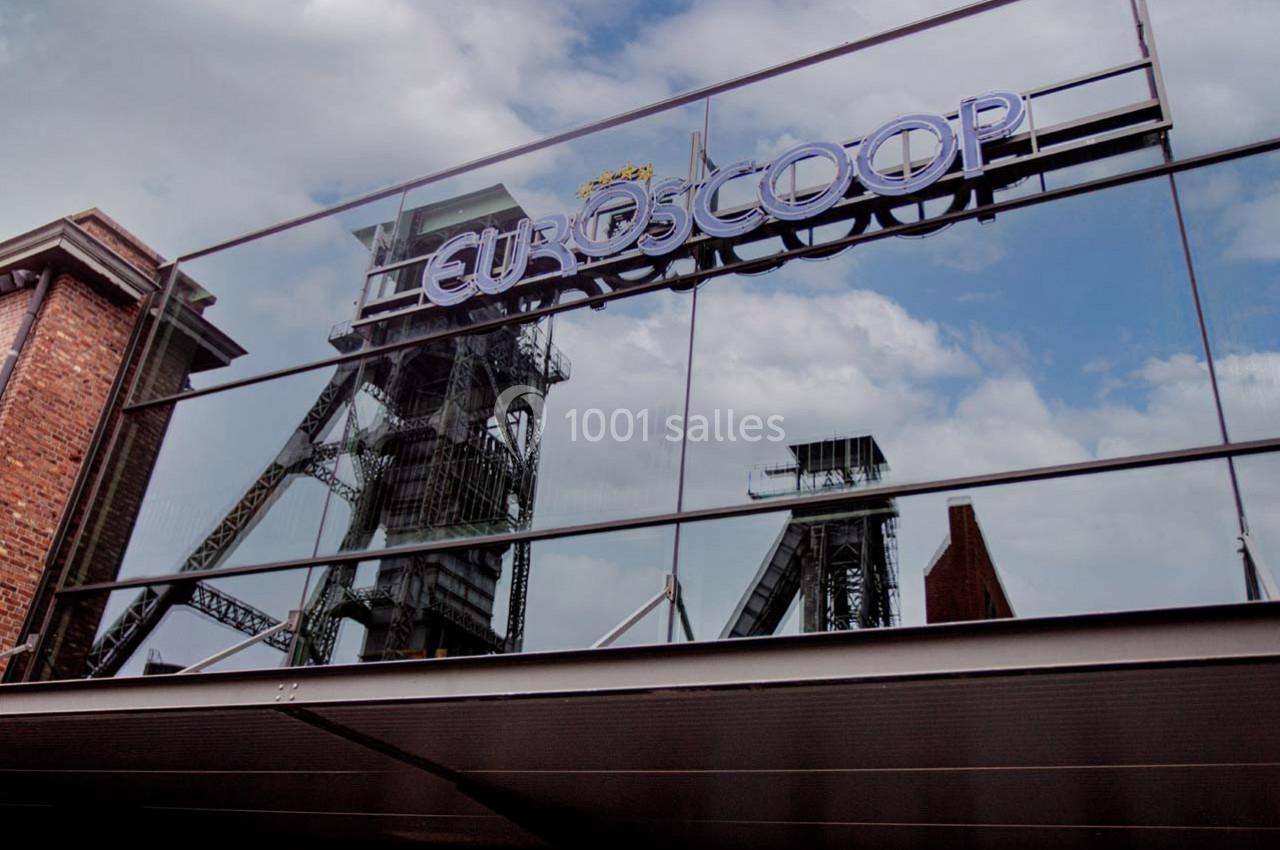 Reflet d'une structure industrielle et d'un ciel nuageux sur la façade vitrée d'un bâtiment avec le logo ’Euroscoop’.