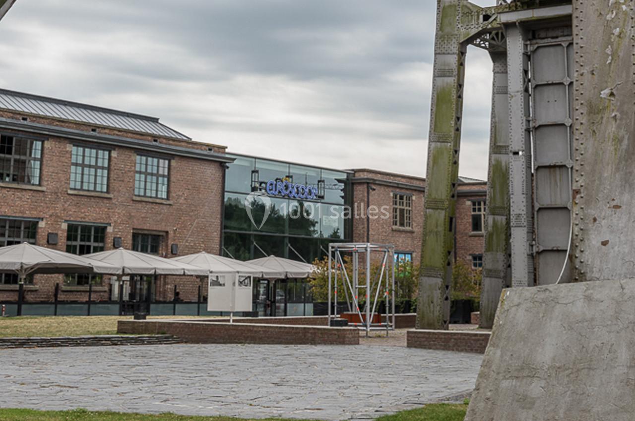 Bâtiments en briques avec grandes fenêtres, terrasse avec parasols et structure métallique au premier plan.