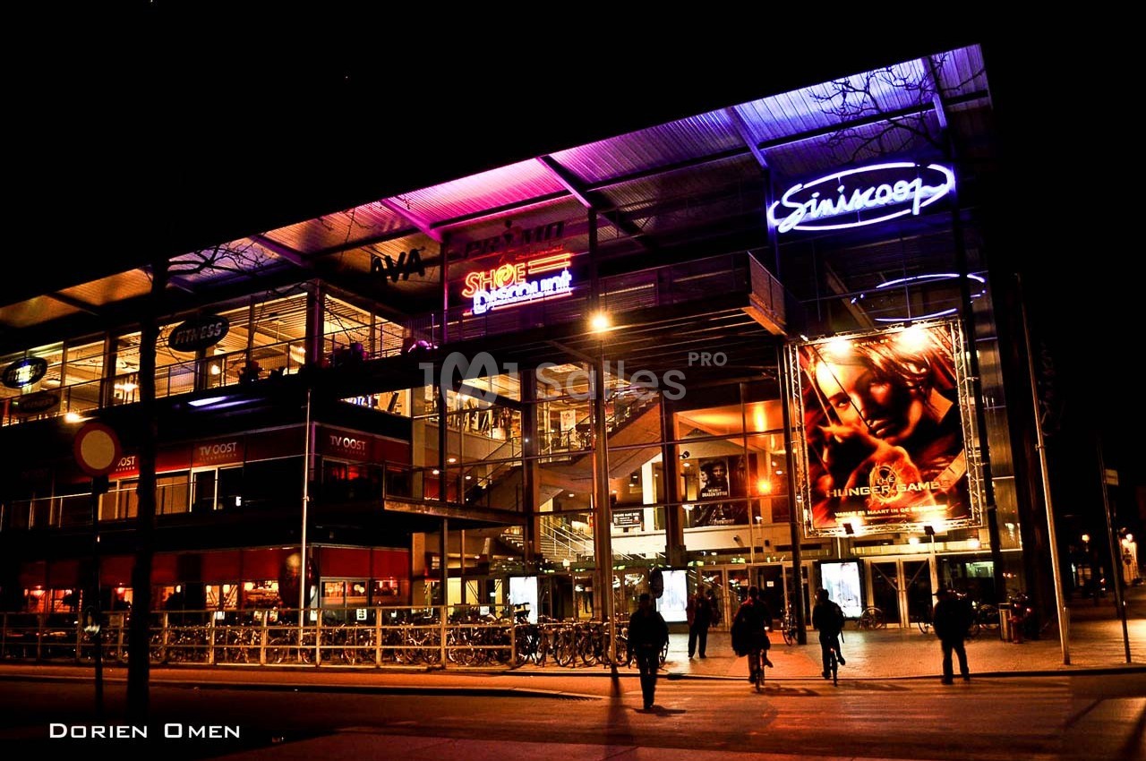 Façade illuminée d'un centre commercial avec enseignes lumineuses et vélos stationnés devant, vue de nuit.