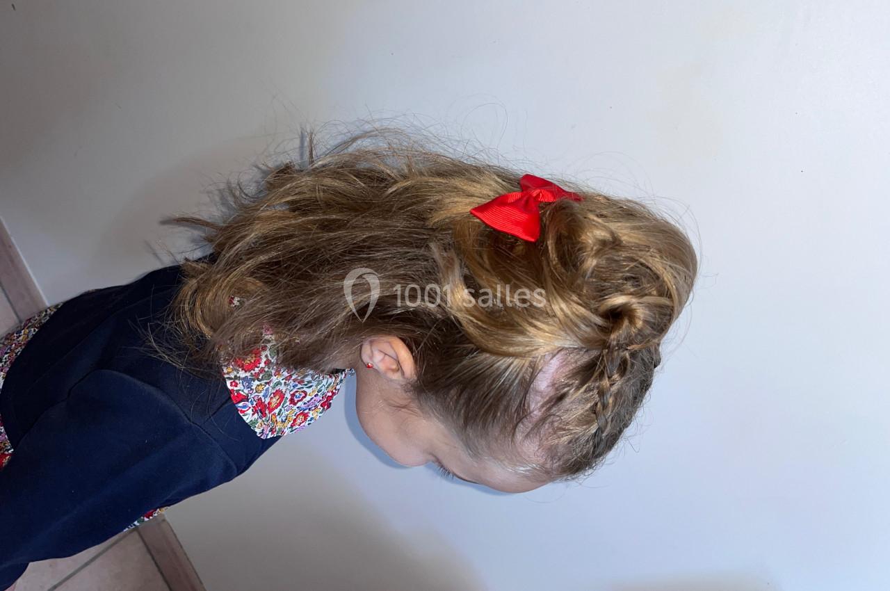 Fillette de profil avec une coiffure tressée ornée d'un nœud rouge, debout devant un mur clair.