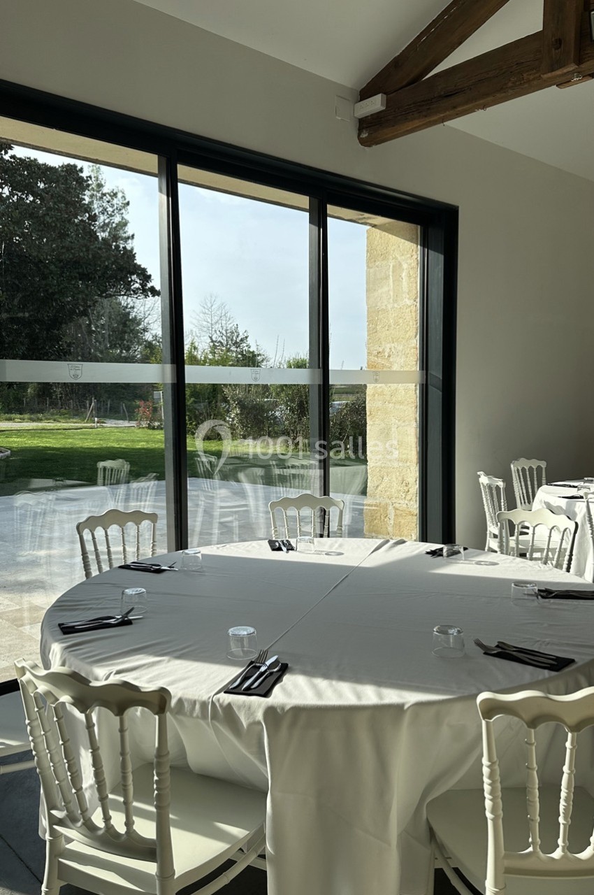 Salle lumineuse avec tables rondes dressées, nappes blanches, chaises élégantes et vue sur un jardin à travers de grandes…