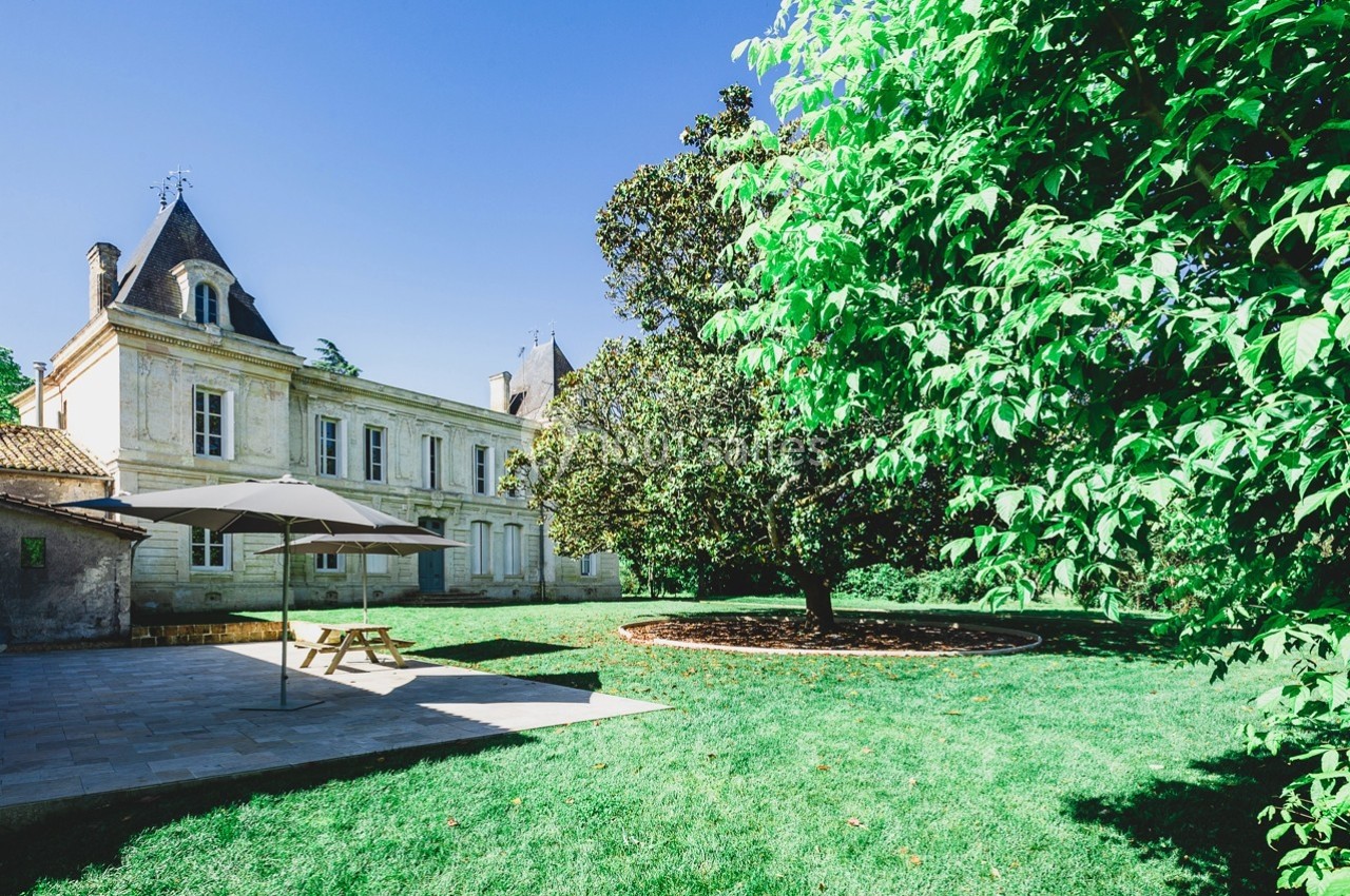 Manoir en pierre avec jardin verdoyant, terrasse ombragée et arbres sous un ciel bleu clair.