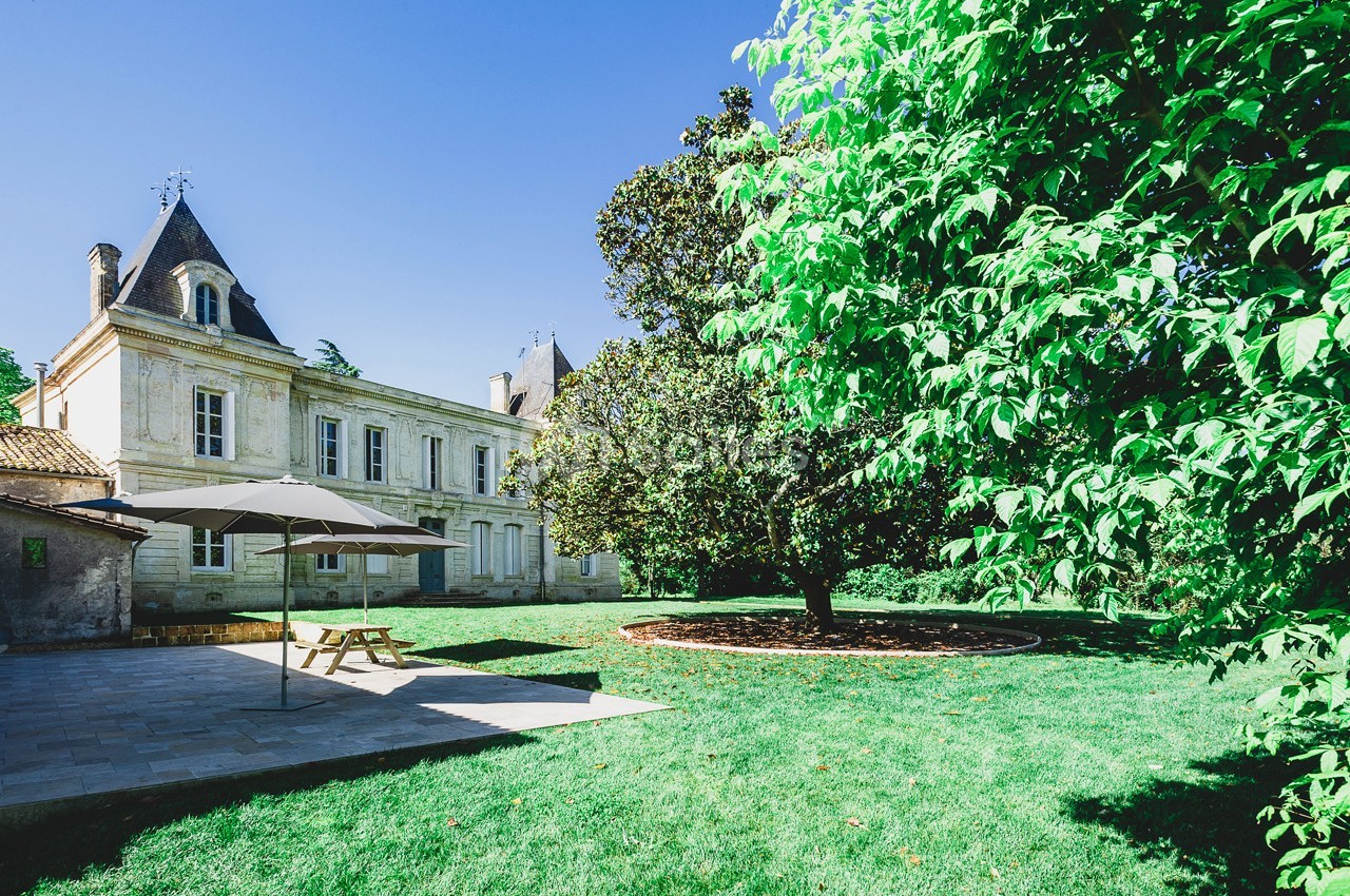 Manoir en pierre avec jardin verdoyant, terrasse ombragée et arbres sous un ciel bleu clair.