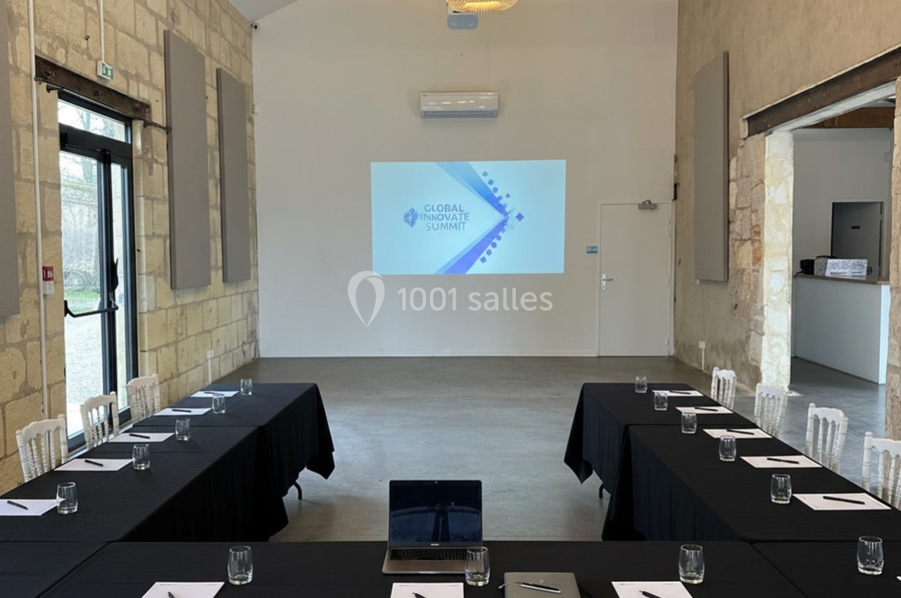 Salle de réunion lumineuse avec tables disposées en U, écran de projection allumé et matériel de prise de notes.